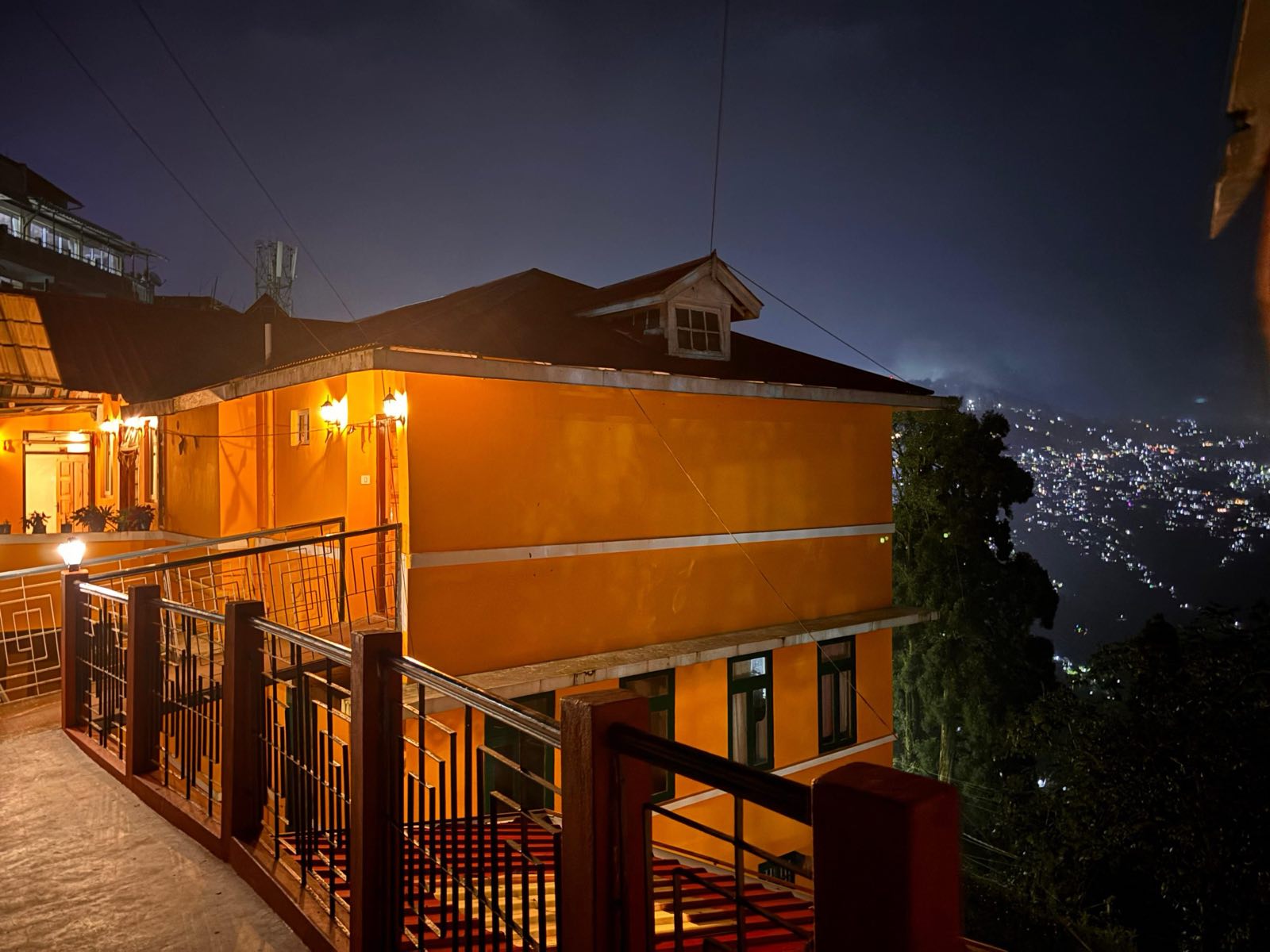 Asraya's saffron-painted cottage lit from within at dusk, with the lights of Darjeeling town scattered on the hillside below